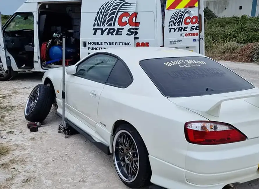 A white sports car is being serviced for a tire change by a Mobile Tyre Fitting Weymouth van on a gravel surface.
