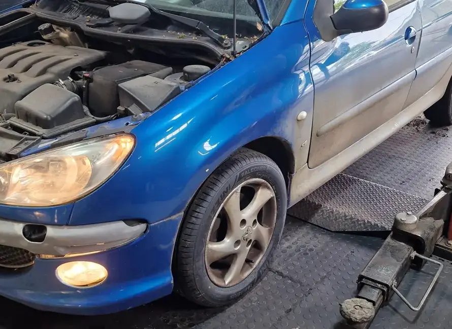 A blue car with its hood open is parked in a garage, ready for Car Battery Fitting Weymouth, with a car jack near the front wheel and headlights turned on.