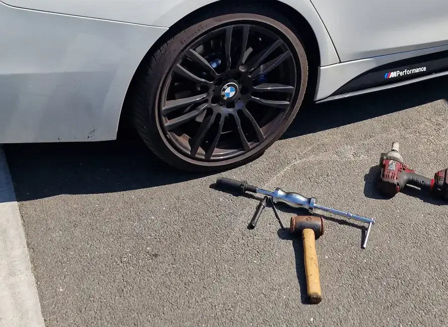 A white BMW with a flat rear tire is parked on asphalt. Nearby, tools for Locking Wheel Nut Removal Weymouth—a hammer, manual tire tool, and cordless impact wrench—are scattered beside the wheel.
