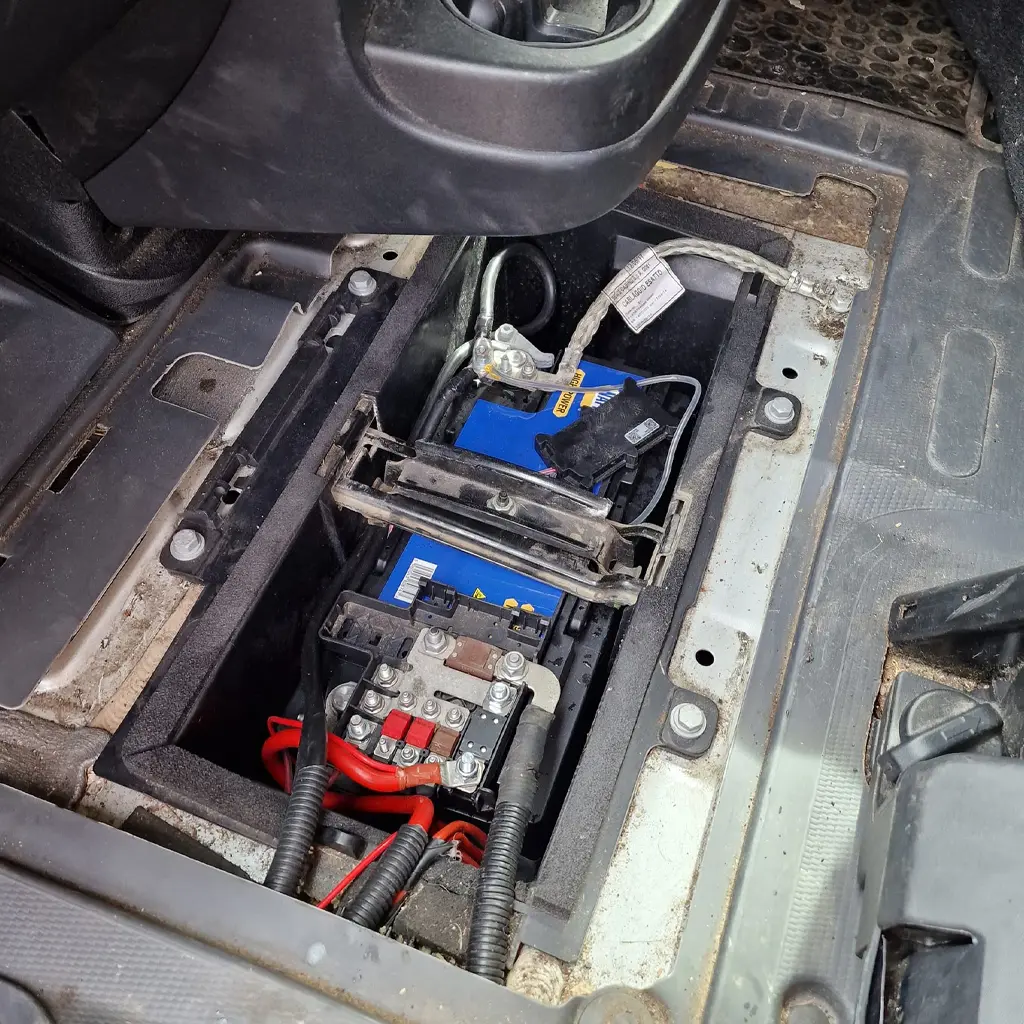A blue car battery and electrical components installed under a vehicle floor panel at Coastal Tyre Services, with wires and connectors visible around the compartment.