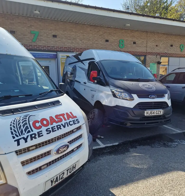 Two Ford service vans, including one labeled "Coastal Tyre Services," are parked outside a building with numbered green doors. The scene is brightly lit by sunlight.