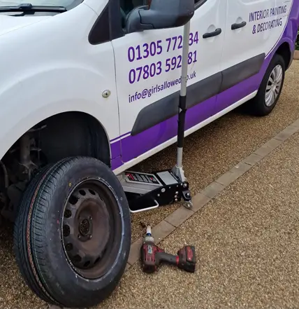 A van with a flat tire is being repaired by Coastal Tyre Services; the front wheel is removed, a jack lifts the vehicle, and a power tool lies beside the spare tire on the ground.