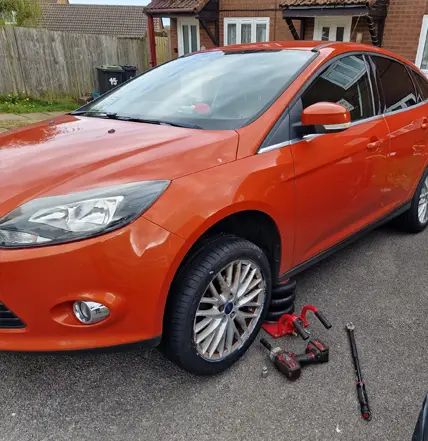 An orange car parked on a driveway with the front left wheel removed; tools and the detached wheel are lying on the ground nearby, ready for attention from Coastal Tyre Services.