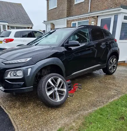 A black SUV with the front left wheel removed is parked on a driveway in front of a house; the removed wheel, serviced by Coastal Tyre Services, lies on the ground nearby.
