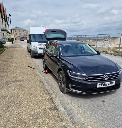 A black Volkswagen with its hood open is parked on the street in front of a white van, adjacent to a construction site and fenced area—possibly awaiting assistance from About Coastal Tyre Services.