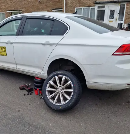 A white sedan with its rear wheel removed is parked on a street; tools and the detached wheel are beside the car, suggesting Coastal Tyre Services might be assisting with a tyre change.